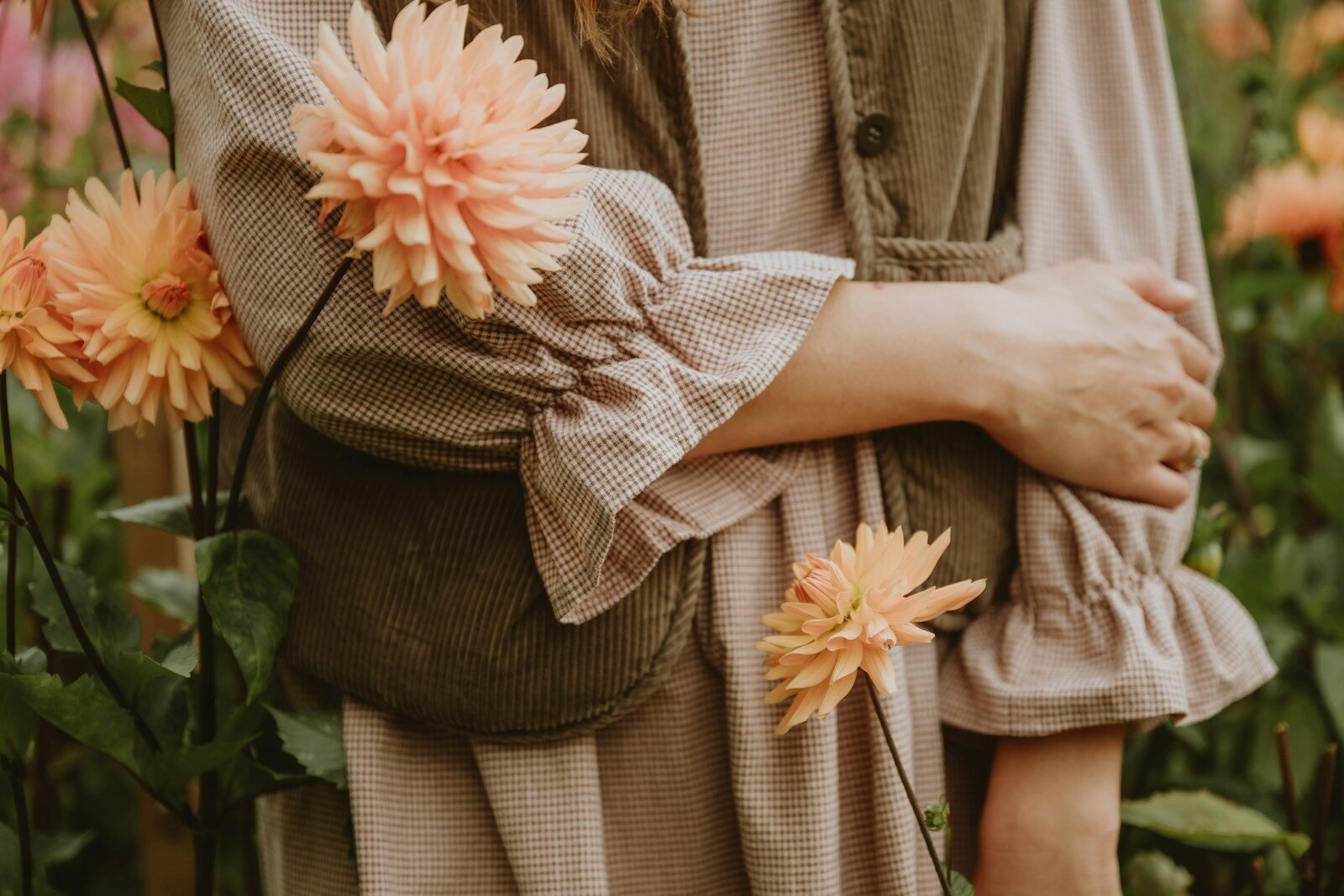 A person in a brown gingham dress and corduroy vest among peach dahlias